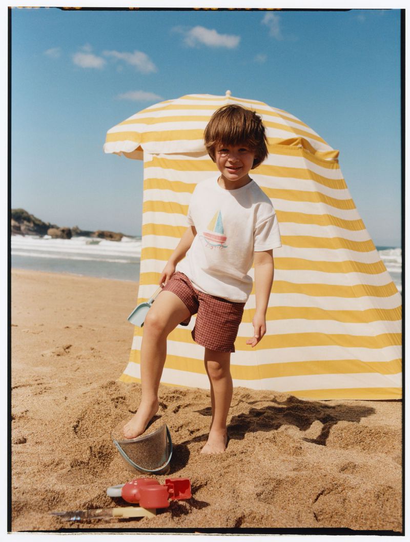 Young child standing on a sandy beach holding a toy shovel in front of a yellow and white striped beach tent.