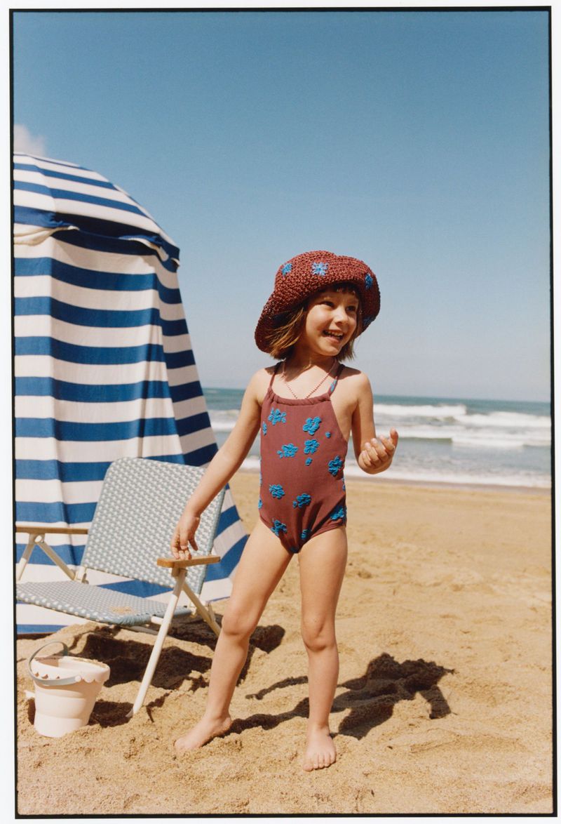 Child wearing brown sun hat and patterned swimsuit on sandy beach beside blue and white striped beach tent