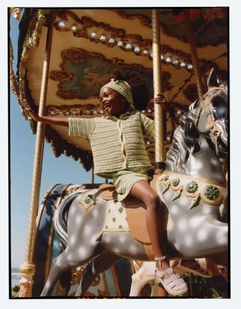 Young child riding a carousel horse wearing a matching knit top, shorts, and headband outdoors in bright sunlight.
