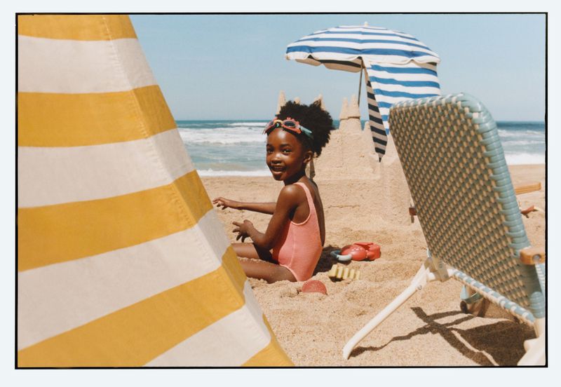 Young child in a pink swimsuit sitting on a sandy beach near a striped beach umbrella and sandcastle
