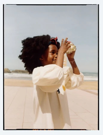 Young child with dark curly hair holding a toy camera against a sandy beach and ocean backdrop for Zara Summer Memories.