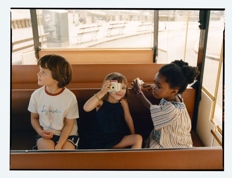 Three children seated on a train bench holding a small camera, photographed by Toby Coulson for Zara.