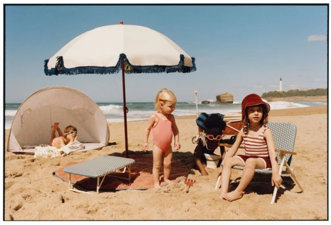 Children playing on a sunny sandy beach with a large parasol, beach chairs, and an ocean backdrop under a clear sky.