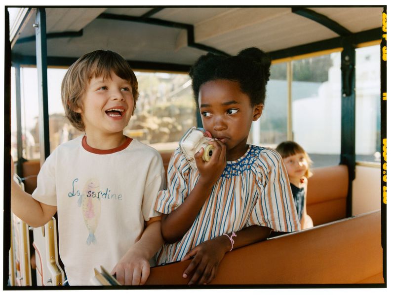 Children riding in a tram, including a boy in a graphic t-shirt and a girl holding a small vintage camera