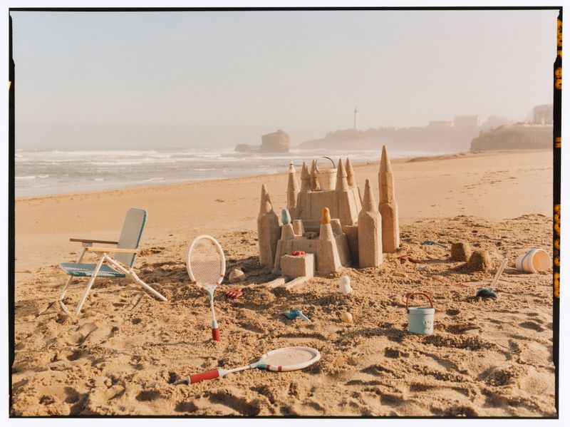 A detailed sandcastle, beach chair, and tennis rackets on a sandy beach by the ocean for Zara Summer Memories.