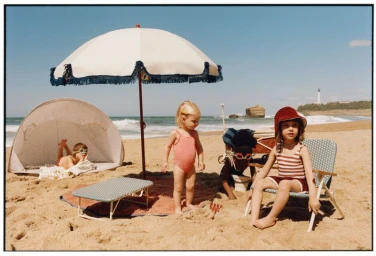 Children playing on a sunny sandy beach with a large parasol, beach chairs, and an ocean backdrop under a clear sky.