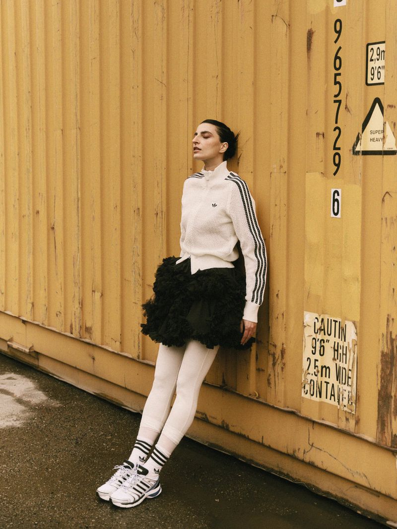 Model leaning against a corrugated yellow shipping container wearing a white track jacket and black ruffled skirt