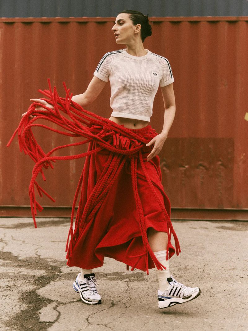 Model wearing a white knit top and red fringed skirt with sneakers against a rusty red container background