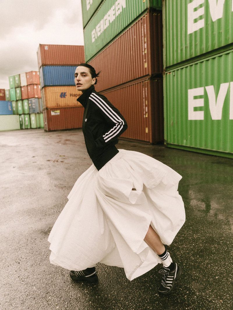 Model in black Adidas track jacket and voluminous white skirt standing before shipping containers