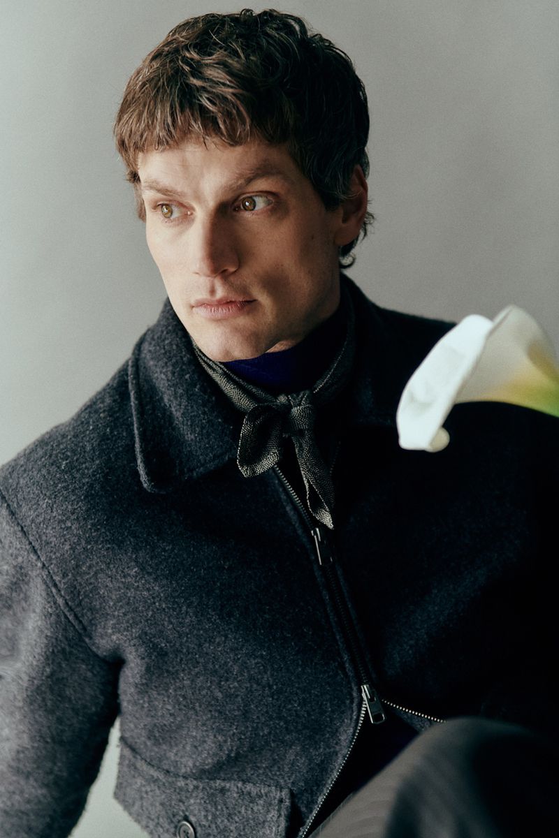 Close-up portrait of a male model wearing a dark wool jacket and a tied scarf, holding a white flower against a neutral grey wall.