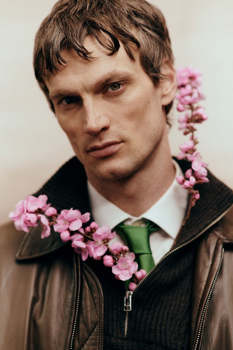 Portrait of a man in a brown leather jacket and green tie with pink flower stems tucked into his collar for Sicky Magazine.