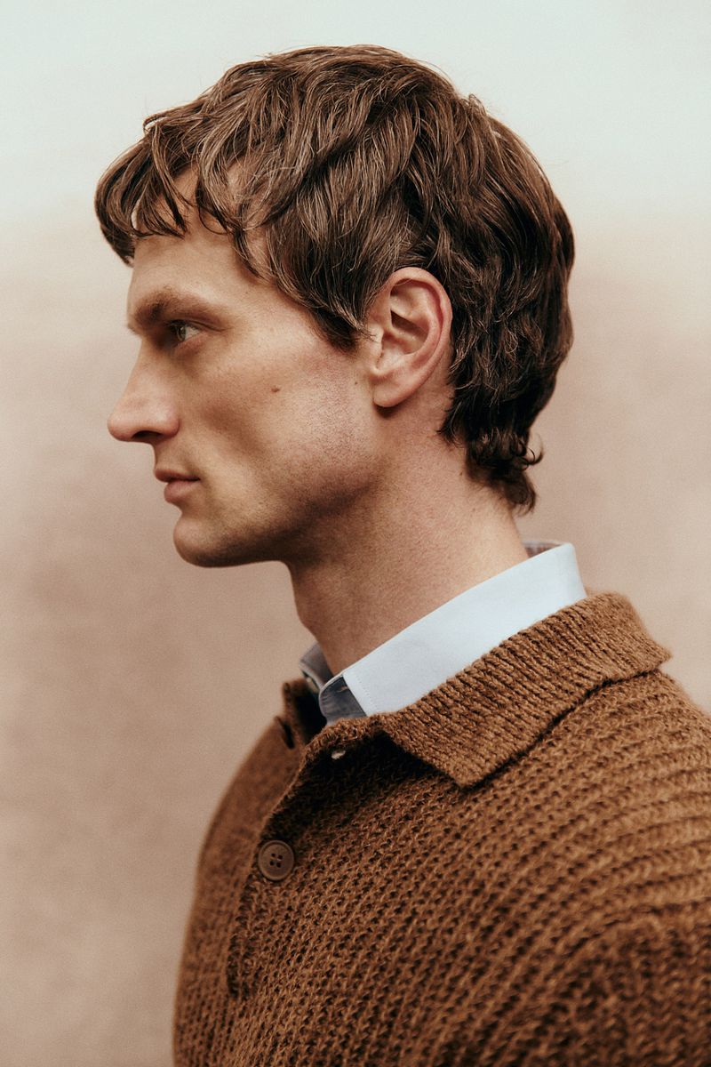 Profile portrait of a fair-skinned adult male with brown hair, wearing a collared shirt and a brown knit cardigan sweater against a neutral backdrop.