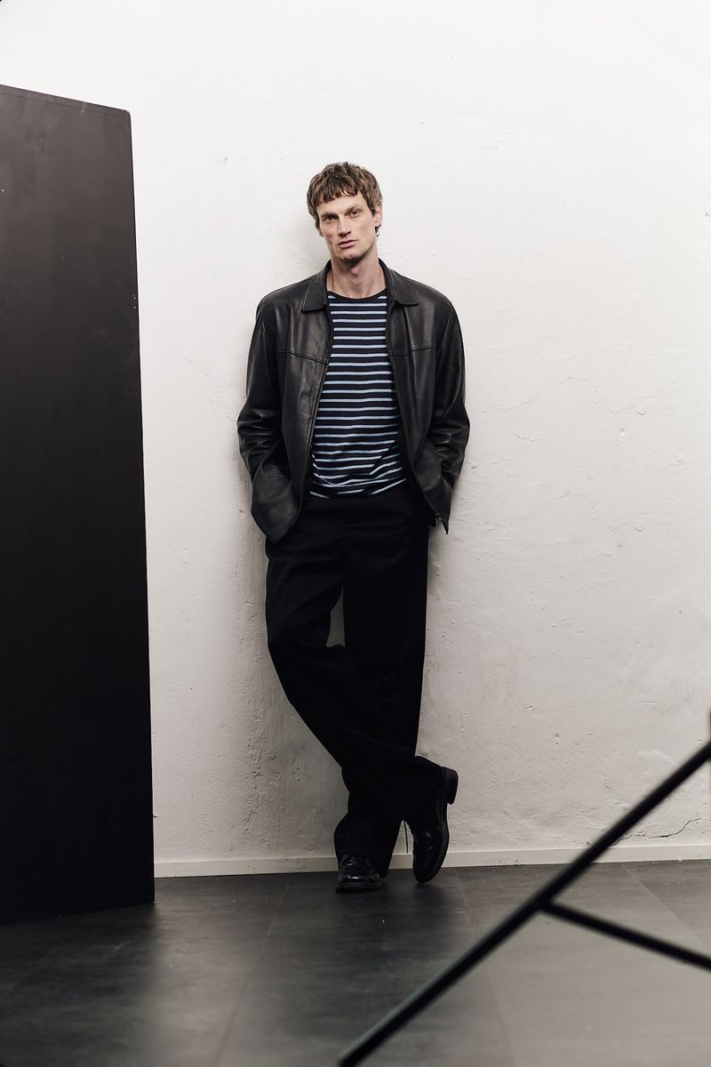 Model leaning against a wall wearing a black leather jacket, striped t-shirt, and dark trousers for Sicky Magazine.