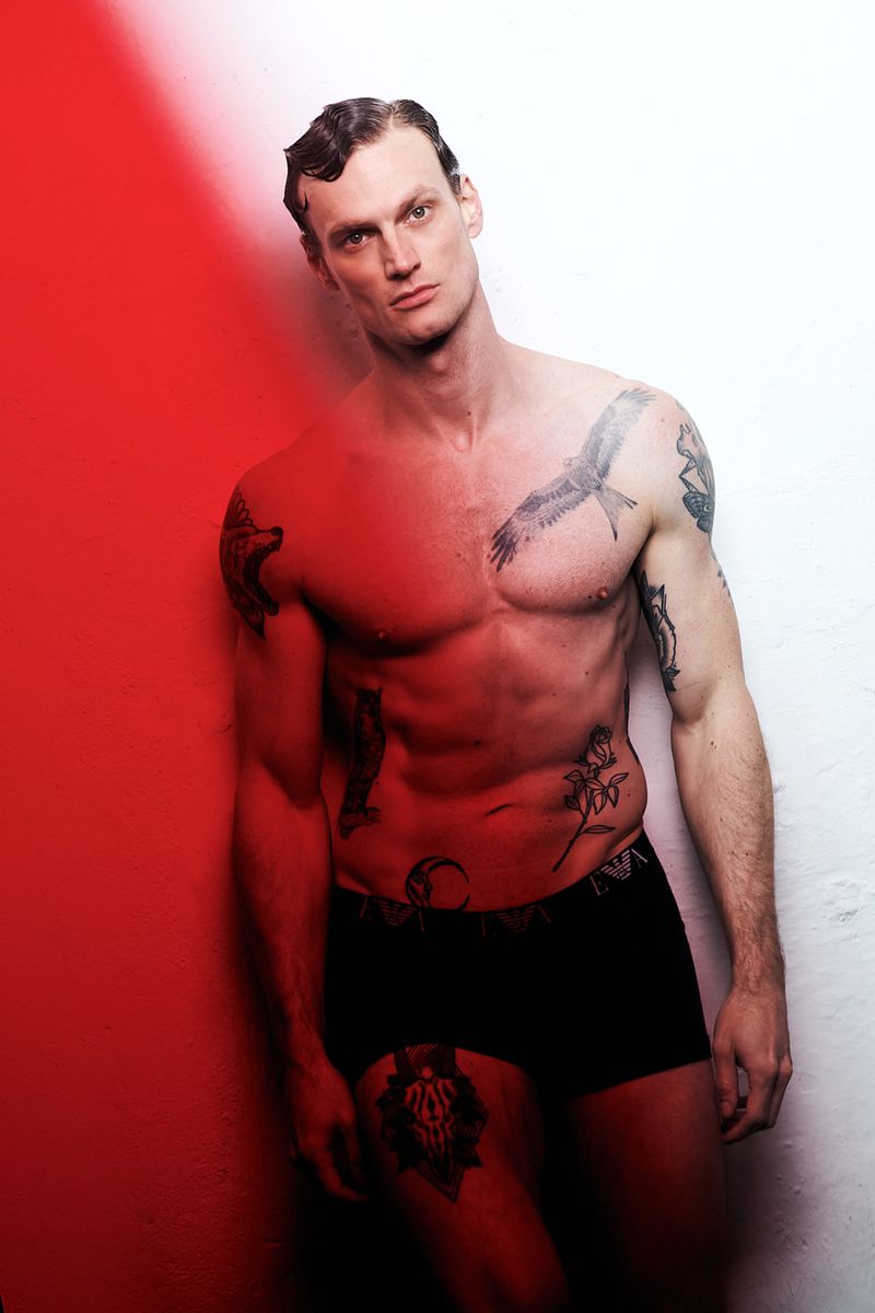 Shirtless tattooed male model wearing black underwear against a red and white background
