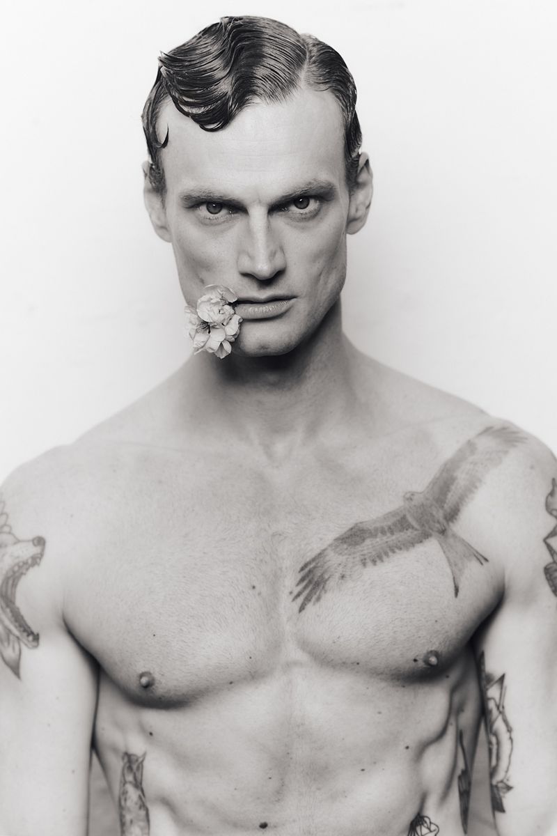 Close-up portrait of a tattooed shirtless man with wet-styled hair holding a flower in his mouth against a light background.
