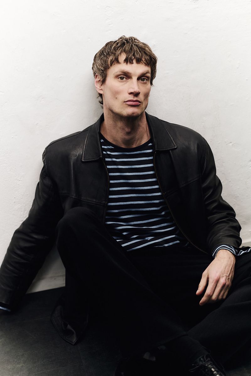Male model wearing a black leather jacket and striped t-shirt seated against a plain white studio wall.