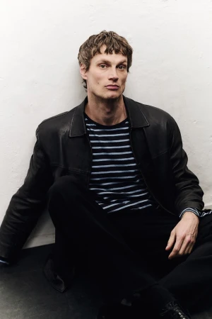 Male model wearing a black leather jacket and striped t-shirt seated against a plain white studio wall.