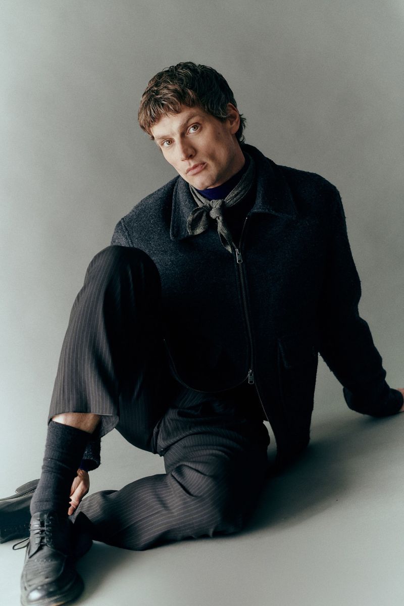 Model seated on grey floor wearing dark wool jacket, trousers, neck scarf, and lace-up shoes for Sicky Magazine editorial.