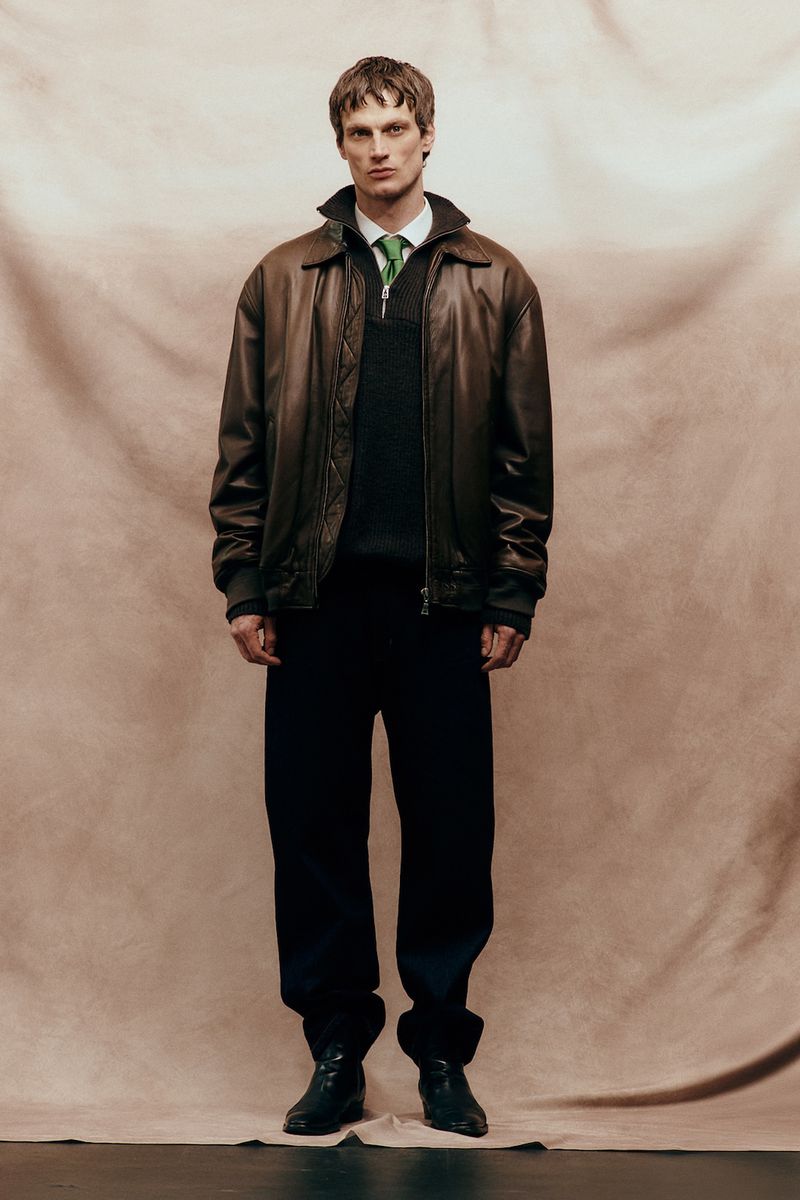 Model Max Bender in brown leather jacket and dark trousers standing against textured fabric backdrop for Sicky Magazine.