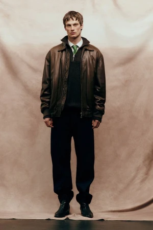 Model Max Bender in brown leather jacket and dark trousers standing against textured fabric backdrop for Sicky Magazine.