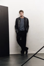 Model leaning against a wall wearing a black leather jacket, striped t-shirt, and dark trousers for Sicky Magazine.
