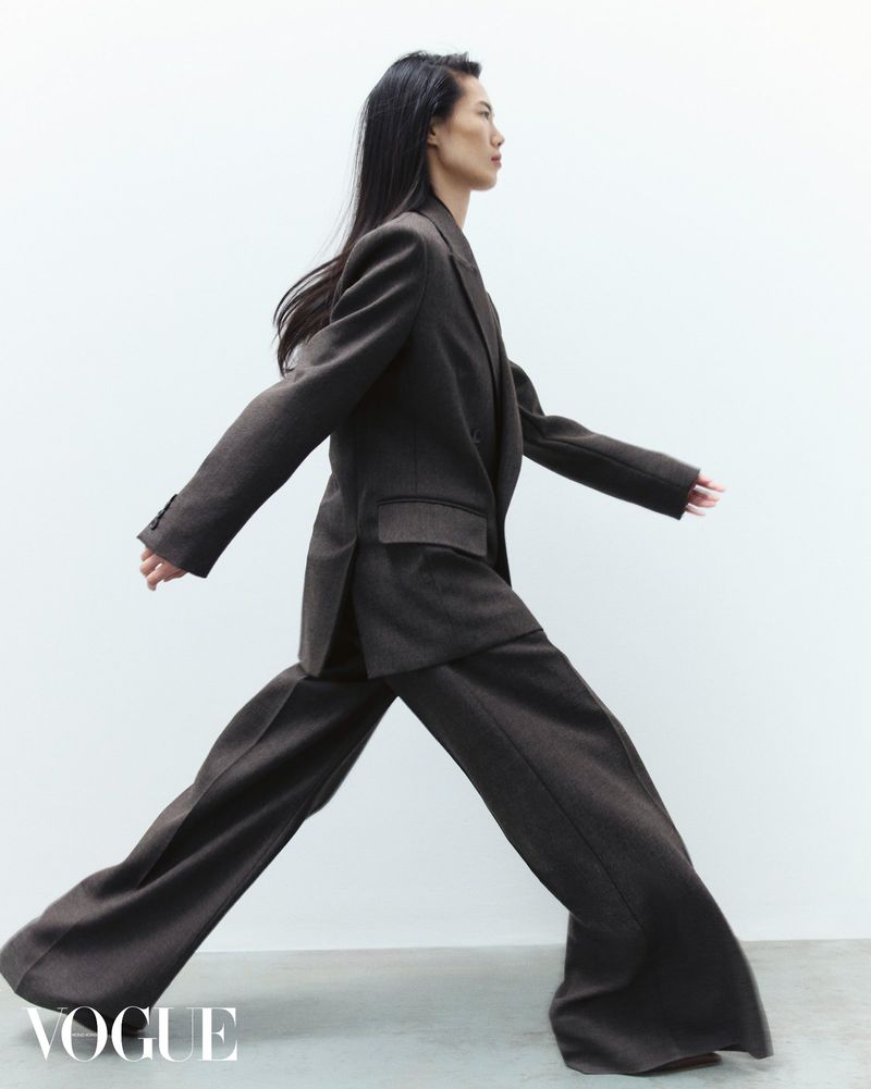 Model with long dark hair walking in profile wearing a dark brown suit against a white background