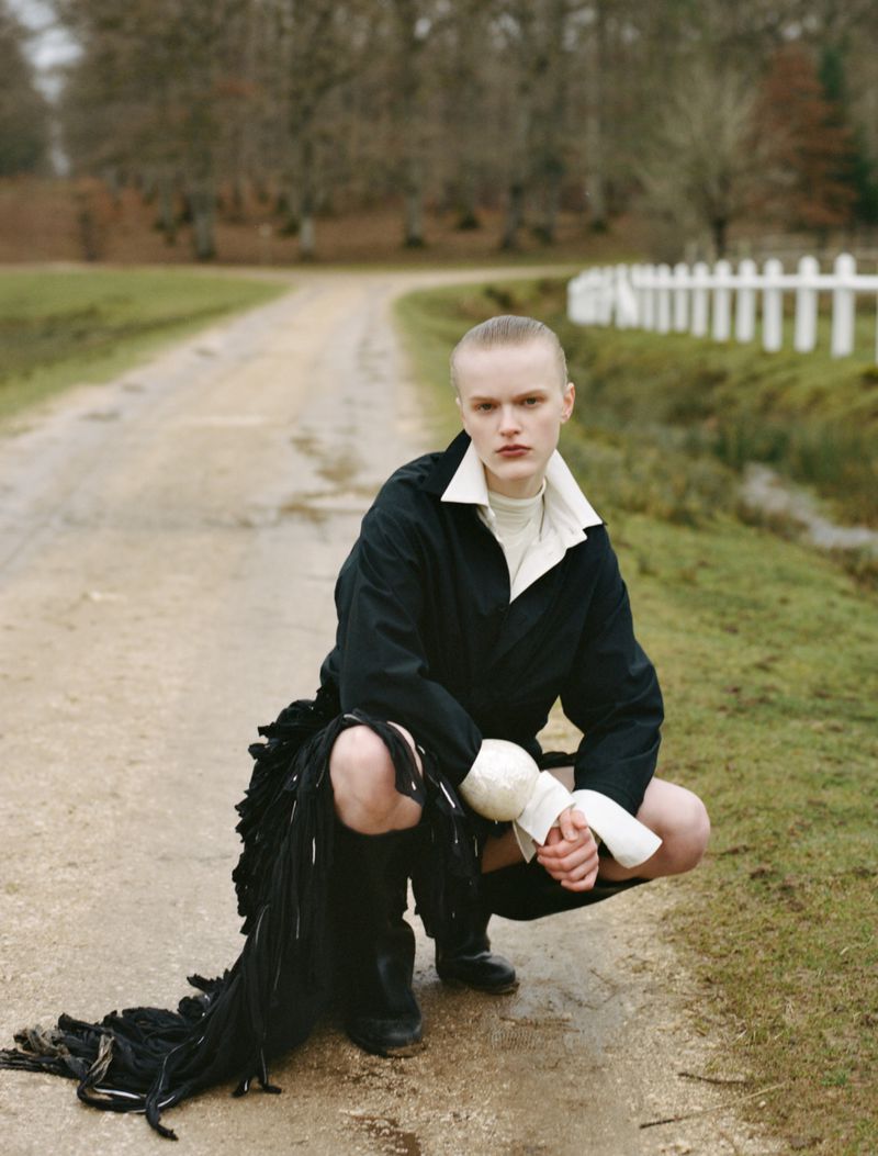 Model crouching on an outdoor dirt path wearing a dark jacket with white collar and black boots
