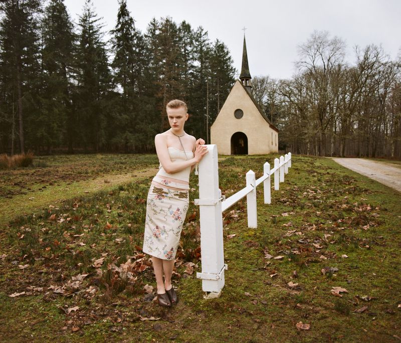 Model standing outdoors near a white wooden fence with a small church and forest in the background