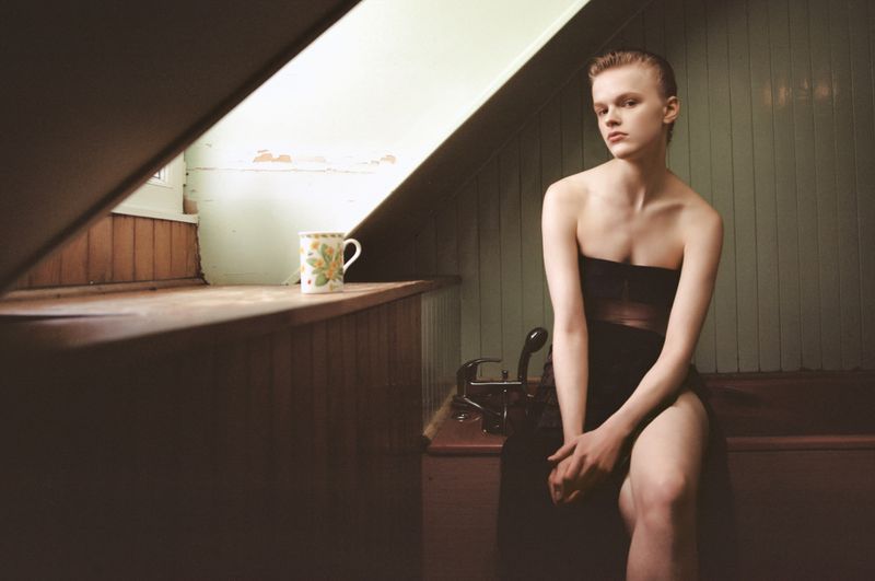 Model wearing a dark strapless dress seated inside an attic bathroom next to a sloping window and patterned mug