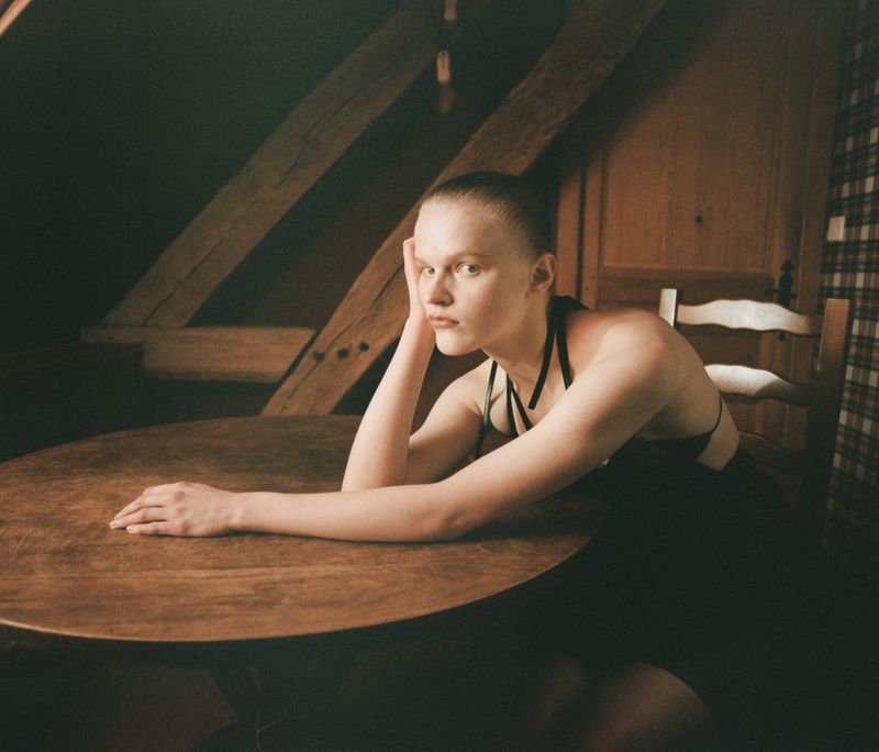 Model Hannah Motler seated at a wooden table in an attic room, resting her head on her hand for Nothing's gonna hurt you editorial.