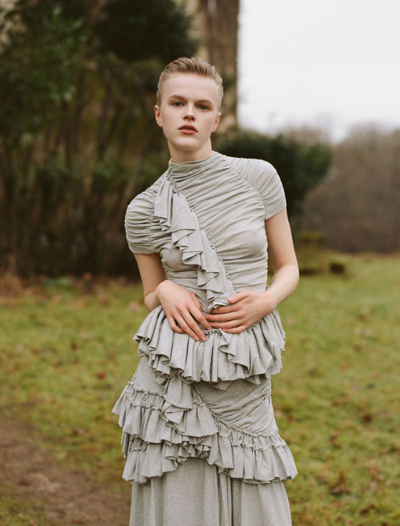 Model Hannah Motler wearing a ruffled grey dress standing outdoors in a natural landscape for editorial