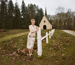Model standing outdoors near a white wooden fence with a small church and forest in the background