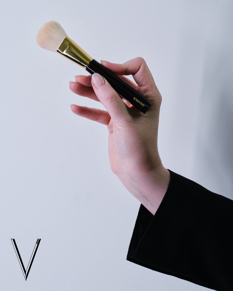 Adult female hand holding a black and gold makeup brush against a neutral grey background in a beauty or fashion context.
