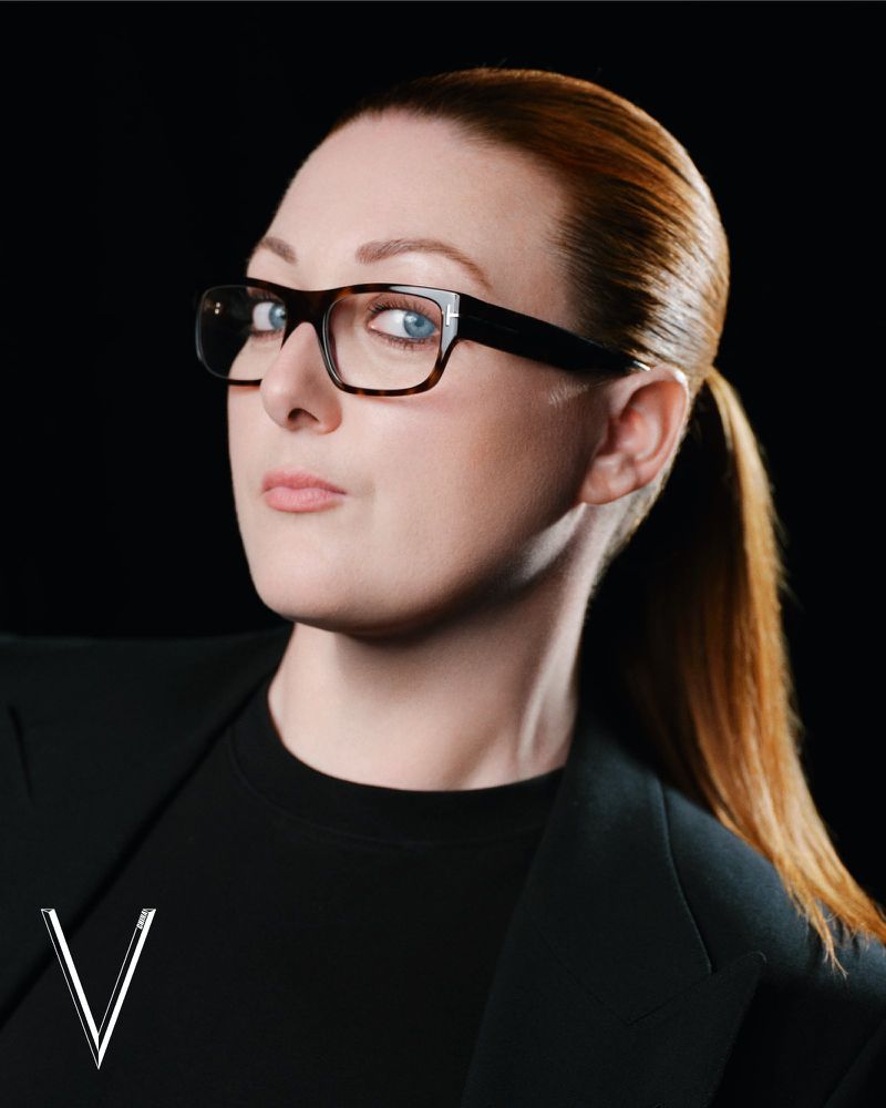 Portrait of a woman with auburn hair in a ponytail wearing tortoiseshell glasses and a black jacket against a dark background.