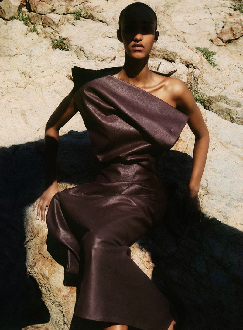Model Muna Mahamed wearing a dark brown structured dress seated on a rocky cliff face for Vanity Fair Italia