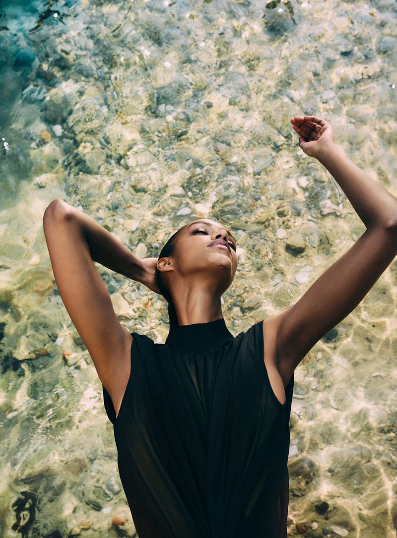Model wearing a black sleeveless top with arms raised against clear shallow water over a pebble bed