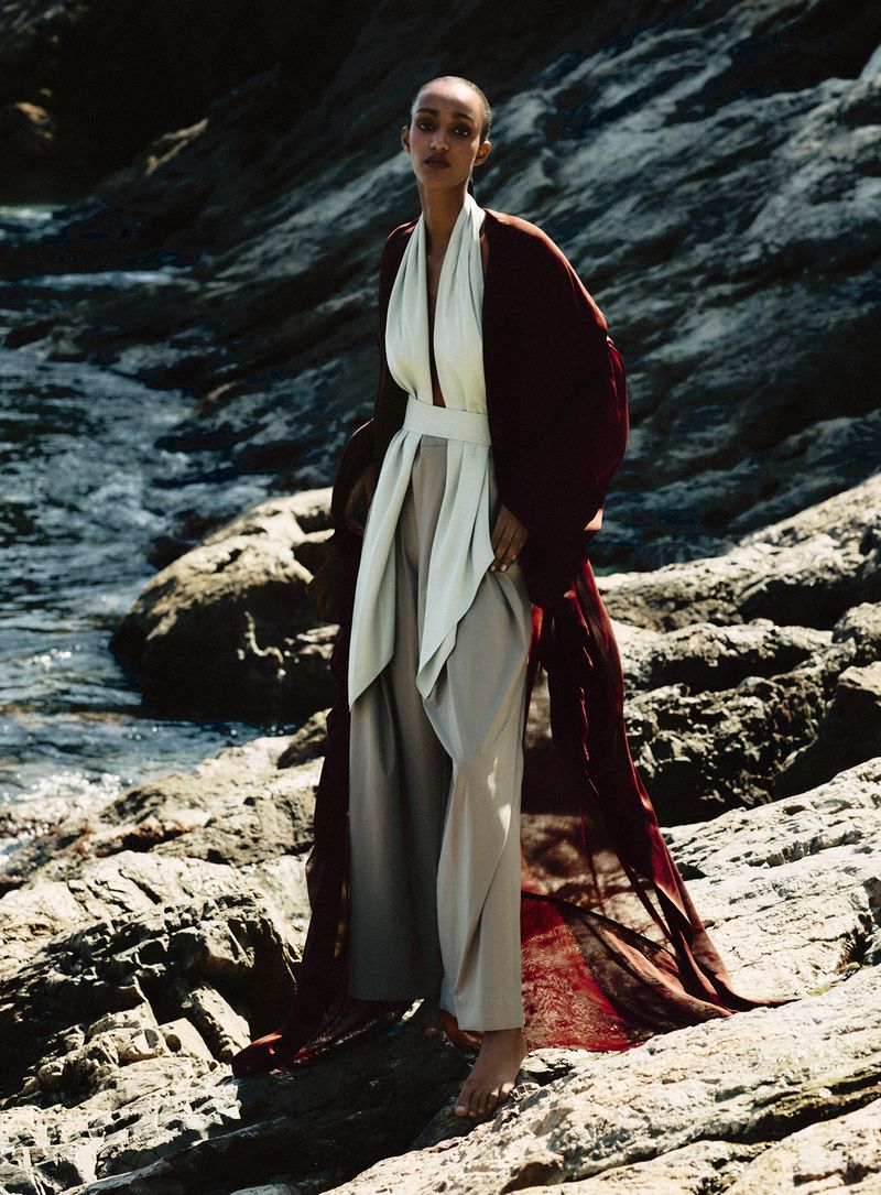 Model Muna Mahamed standing on rocky coastal shore wearing a flowing burgundy robe and pale halter top for Vanity Fair Italia