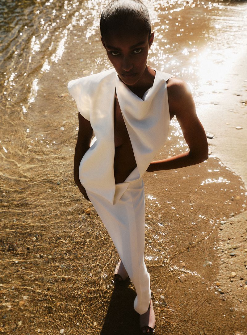 Model standing on a sandy shore by shallow water wearing a structured white dress for a Vanity Fair Italia editorial