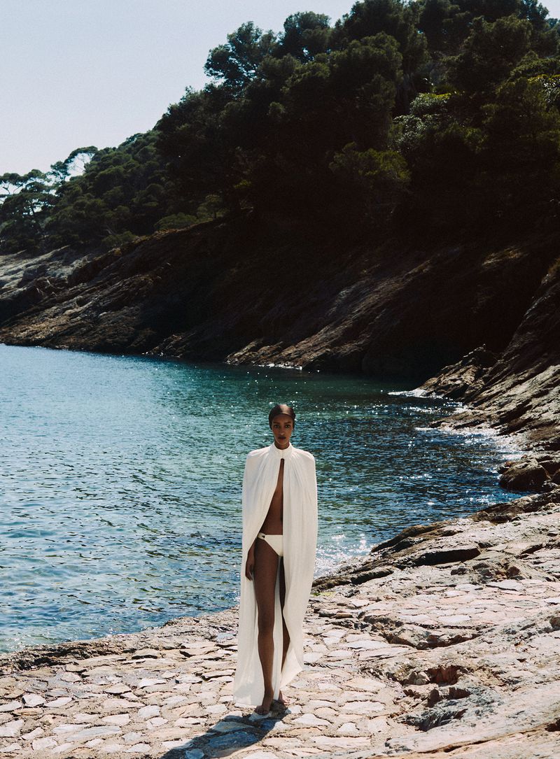 Model standing on a rocky coastal path wearing a long white cape over swimwear with the ocean and forested hillside in the background.