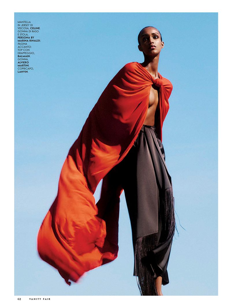 Model in vibrant red cape and dark skirt standing against a clear blue sky for a Vanity Fair Italia fashion editorial.