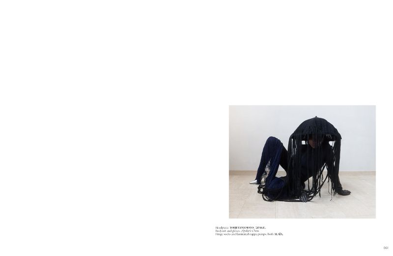 Person in dark clothing and long dark fringe, posed in a partially seated position on a beige surface, against a white backdrop.