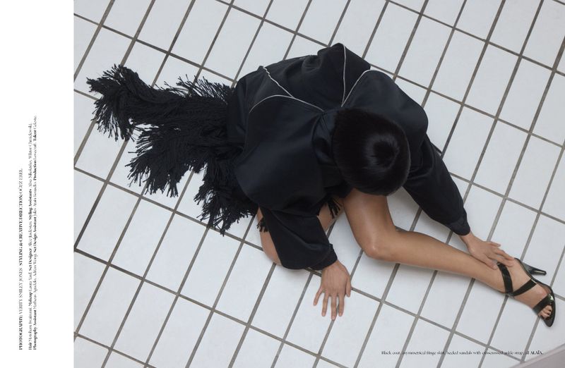 Overhead editorial shot of a woman in a black fringe coat and heeled sandals seated on white tiled floor.
