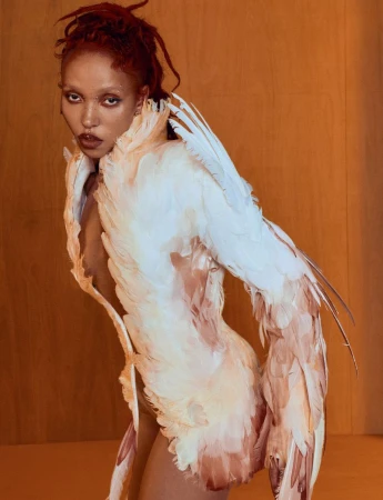 FKA Twigs wearing a sculptural white and peach feathered garment against a warm wood paneled wall.
