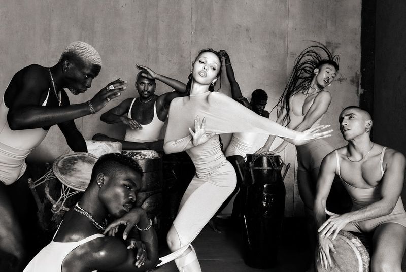 FKA twigs posing centrally in a monochromatic group portrait for 10 Magazine, surrounded by dancers with drums in a concrete studio setting.