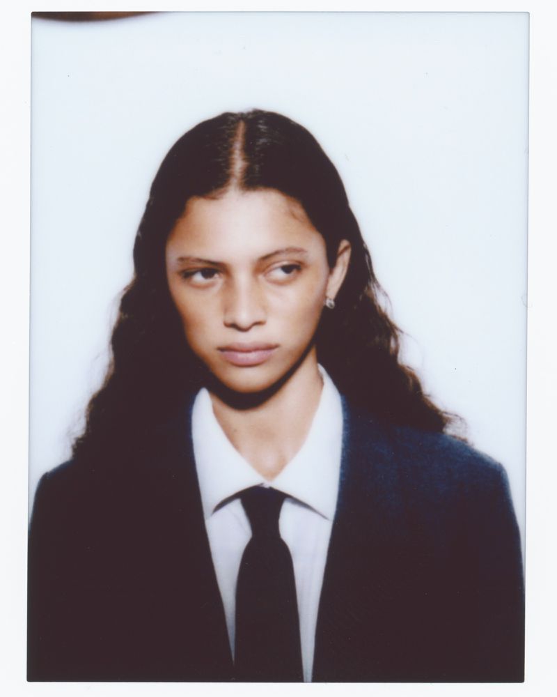 Portrait of a model with long dark hair wearing a dark blazer, white button-up shirt, and dark tie against a white background
