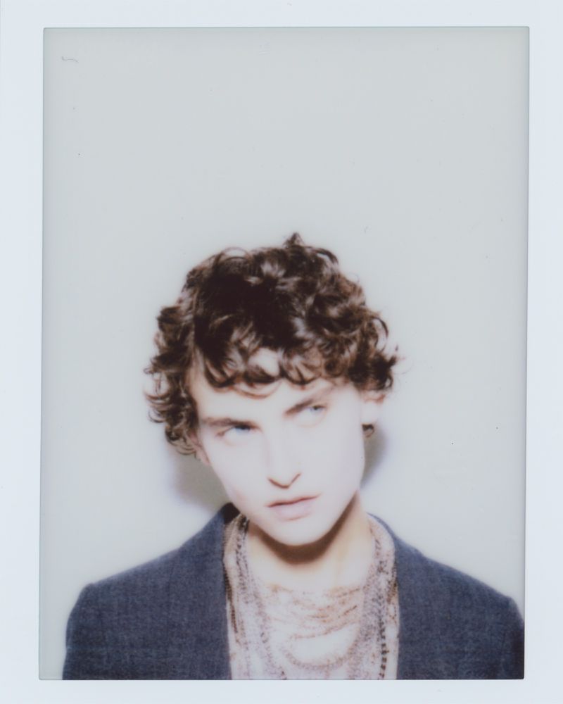 Portrait of a person with curly dark hair wearing a dark blazer and layered necklaces on a white seamless background