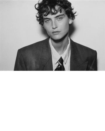 Portrait of a model with curly dark hair wearing a suit jacket, white shirt, and tie against a plain background