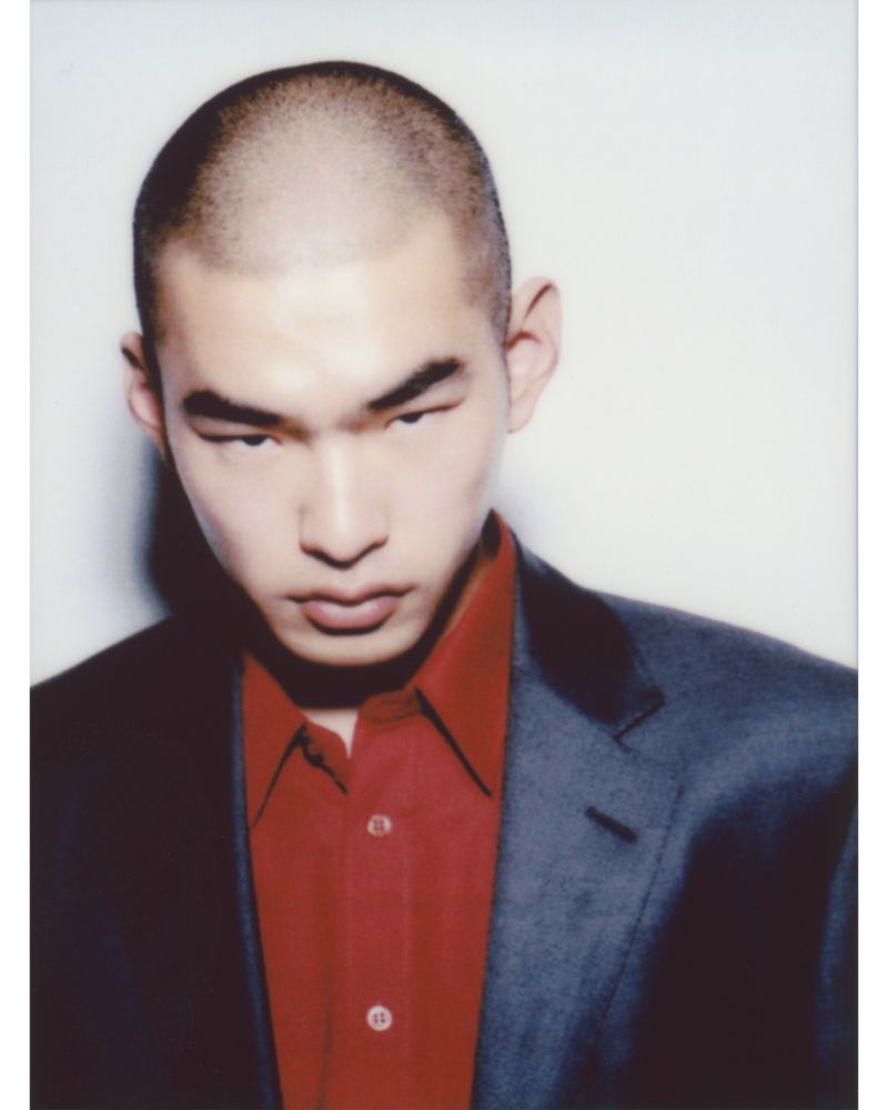 Portrait of an Asian man with a shaved head wearing a red shirt and dark blazer against a white background