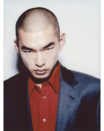 Portrait of an Asian man with a shaved head wearing a red shirt and dark blazer against a white background