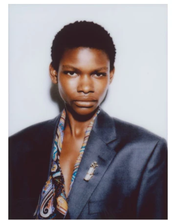 Portrait of a model with short dark hair wearing an oversized grey blazer and patterned scarf against a plain white background